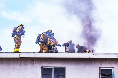 Photo of firemen on top of a roof of a home with smoke coming out of it which could indicate smoke and fire damage.